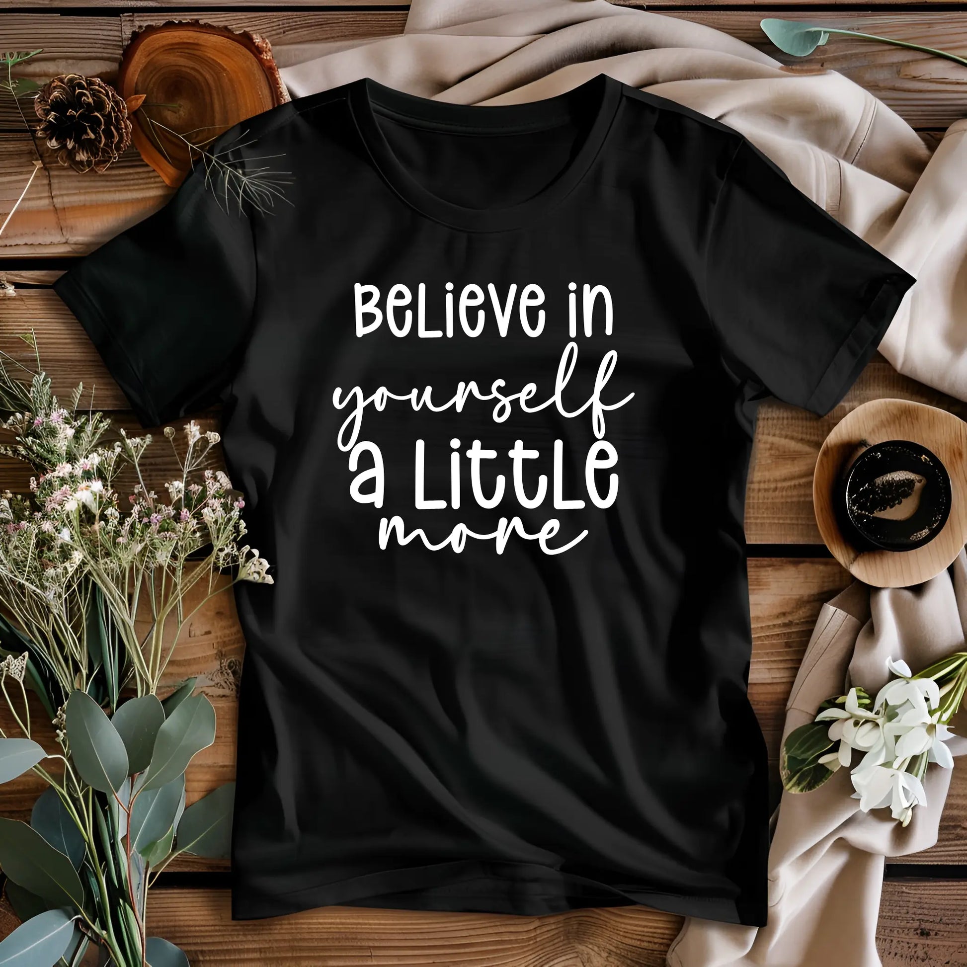 Black t-shirt with 'Believe in yourself a little more' text on a wooden surface with decorative elements.