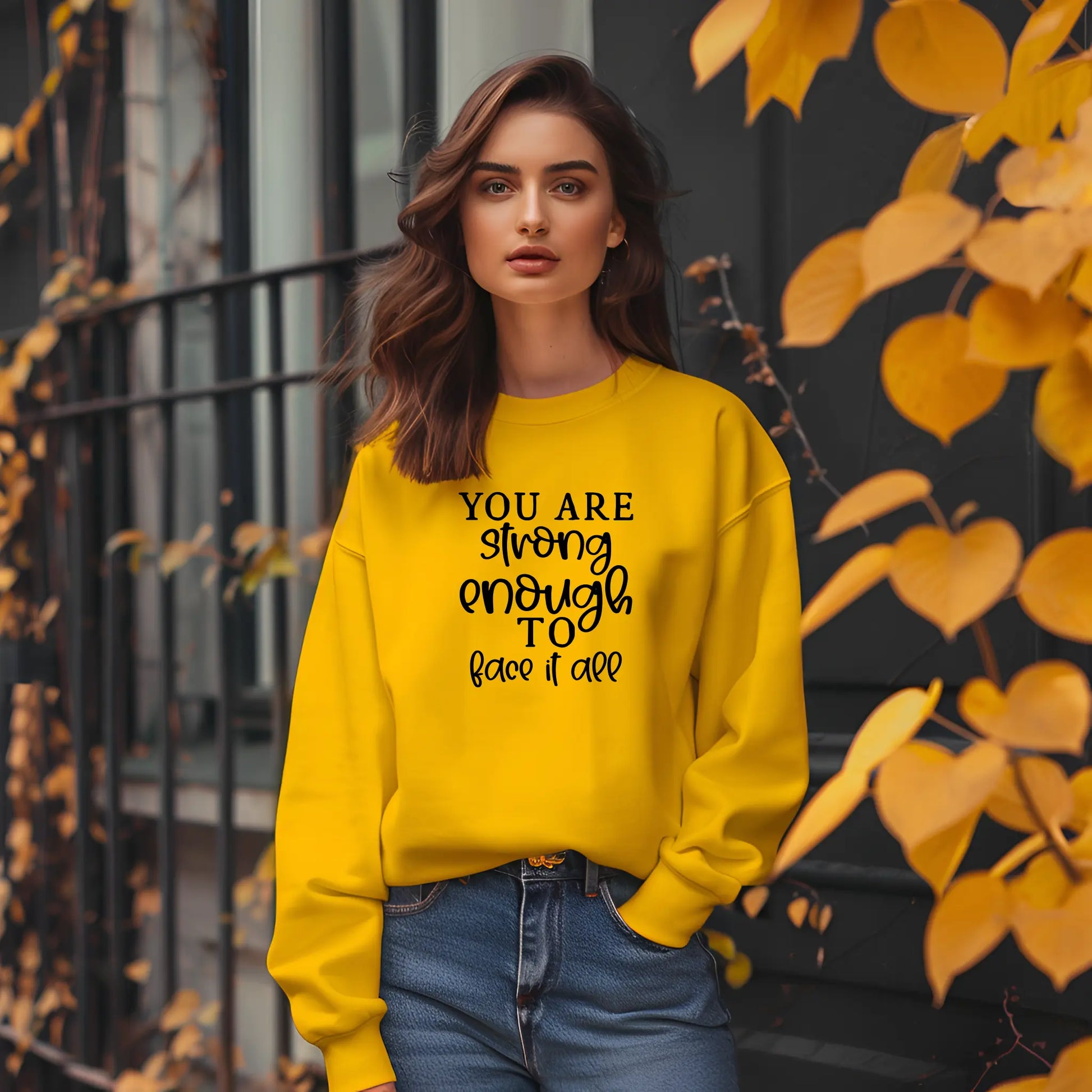 Woman wearing a yellow sweatshirt with an inspirational message, standing in front of autumn leaves.