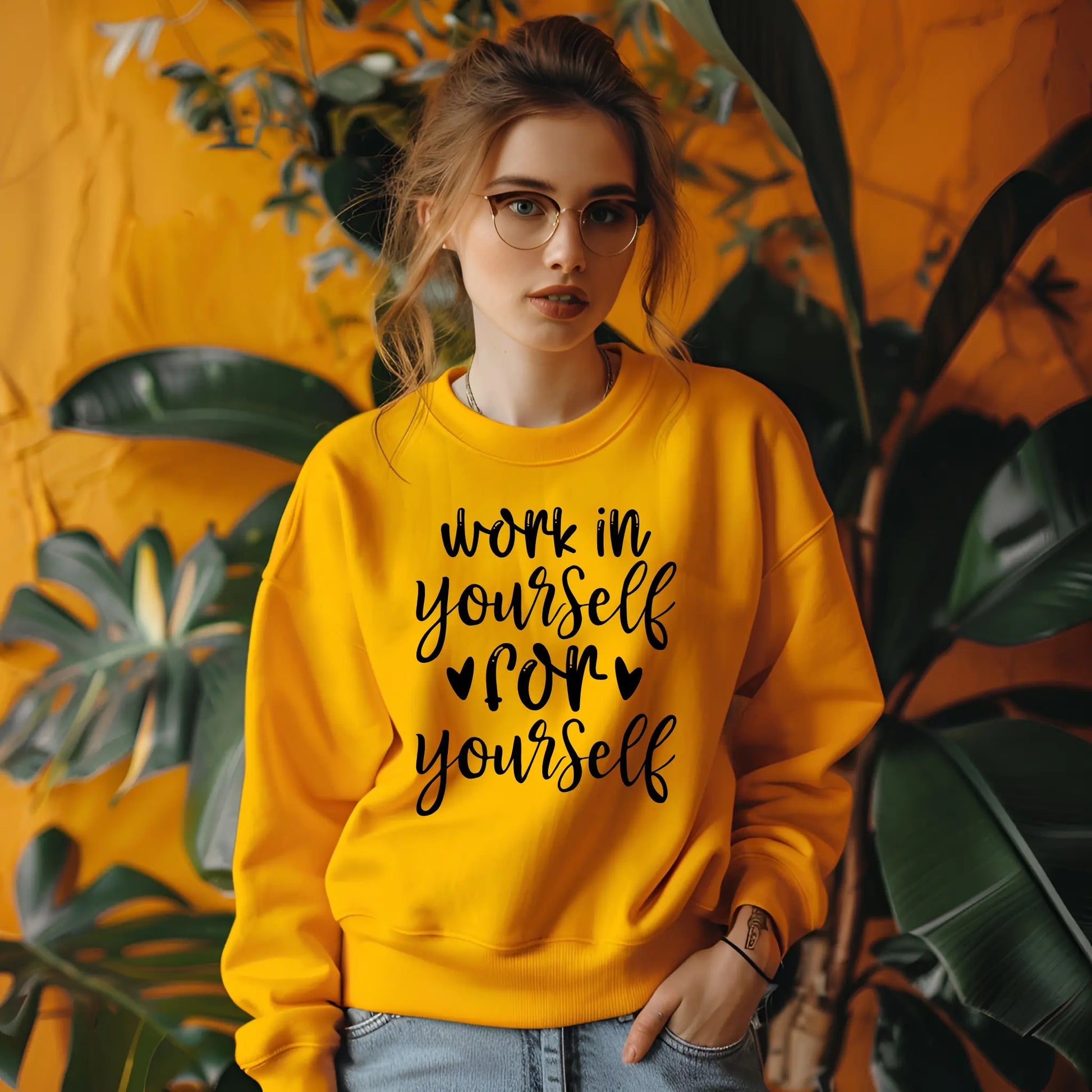 Person wearing a yellow sweatshirt with text, standing against an orange wall with plants.
