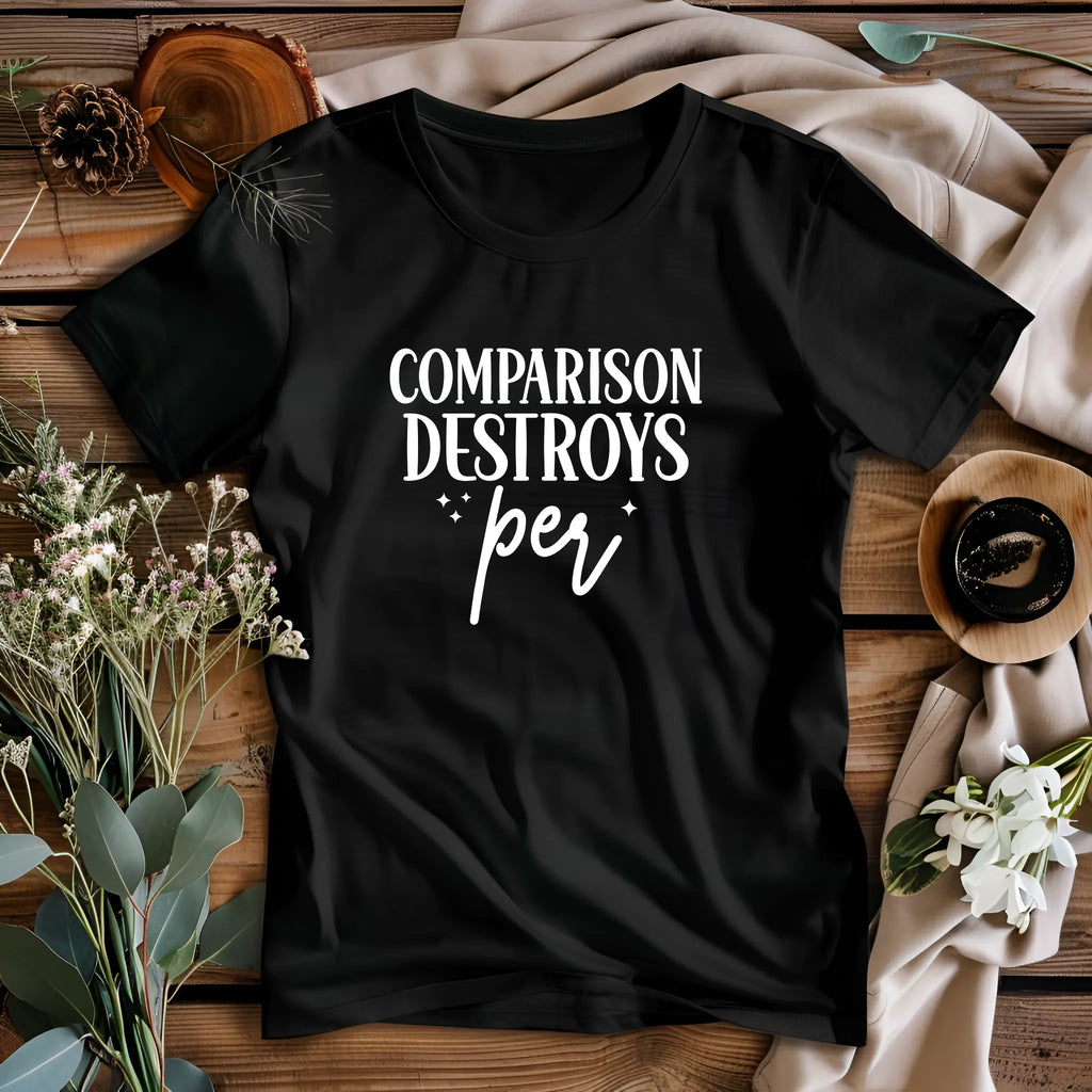 Black t-shirt with 'Comparison Destroys Per' text on a wooden surface with flowers and pinecones.