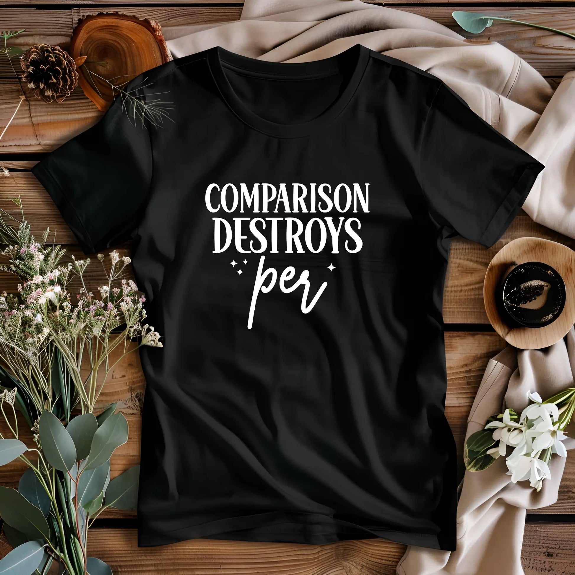 Black t-shirt with 'Comparison Destroys Per' text on a wooden surface with flowers and pinecones.
