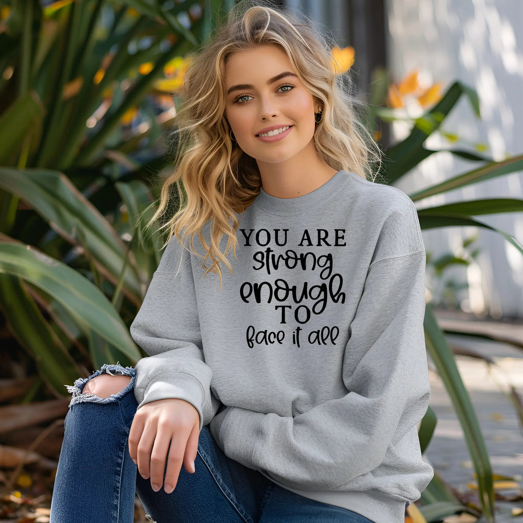 Woman wearing a gray sweatshirt with motivational text, sitting outdoors among plants.