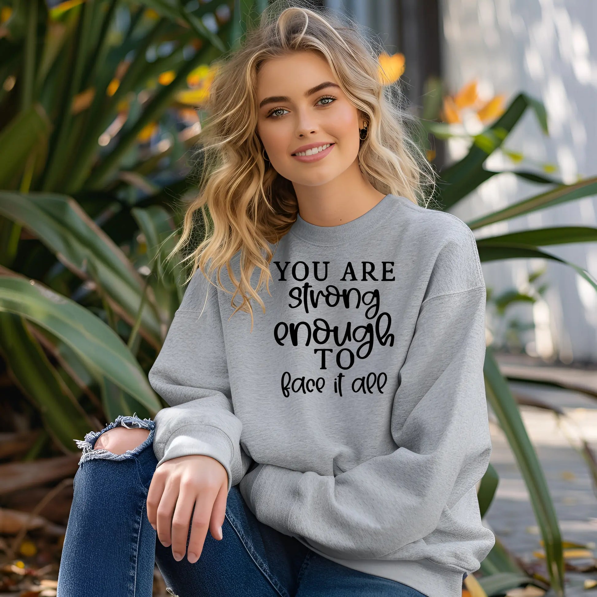 Woman wearing a gray sweatshirt with motivational text, sitting outdoors among plants.