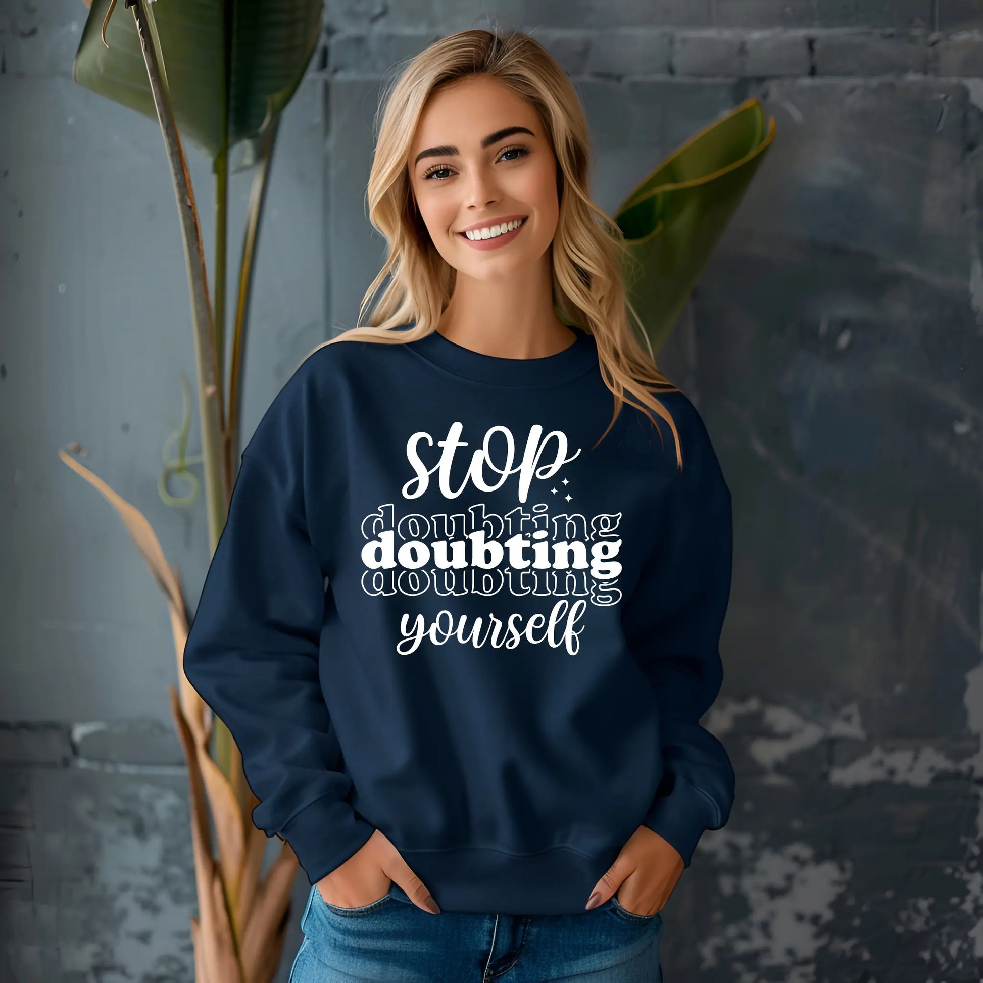 Woman wearing a navy blue sweatshirt with motivational text, standing against a gray wall with plants.
