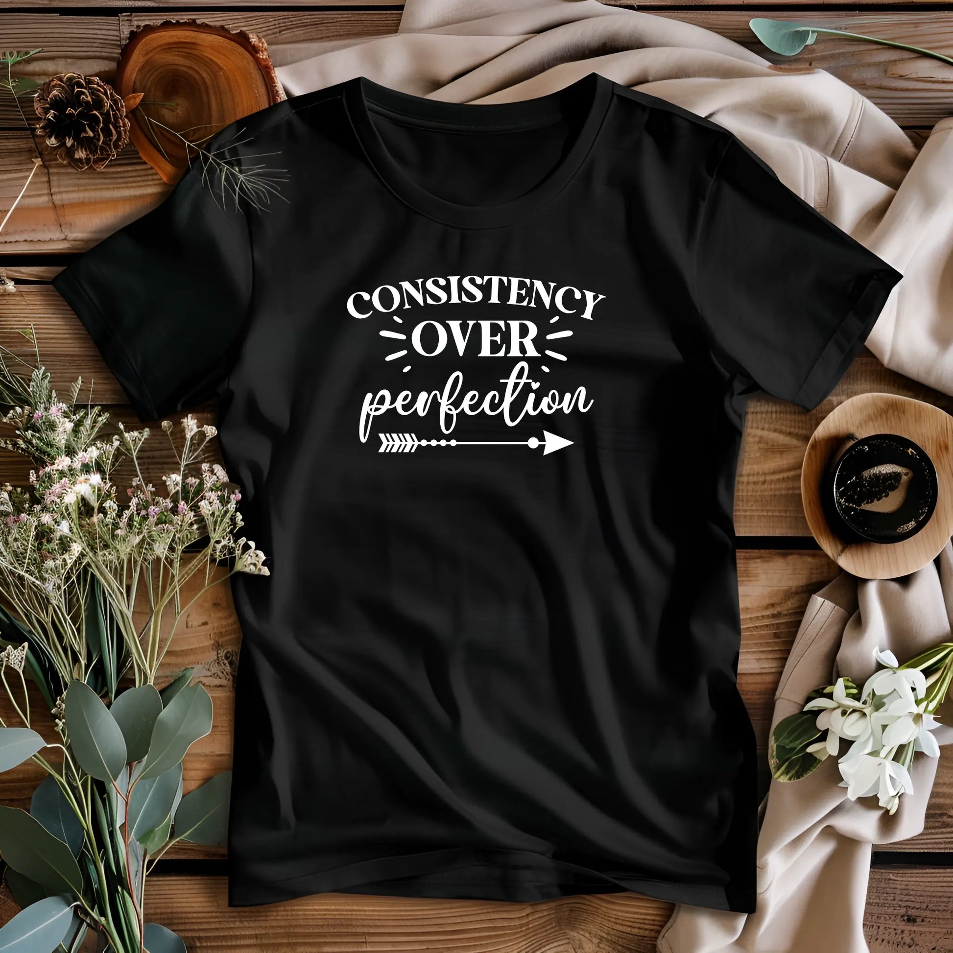 Black t-shirt with 'Consistency over perfection' text on a wooden surface with decorative elements.