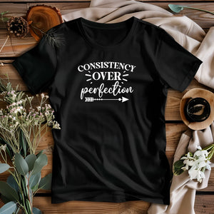 Black t-shirt with 'Consistency over perfection' text on a wooden surface with decorative elements.