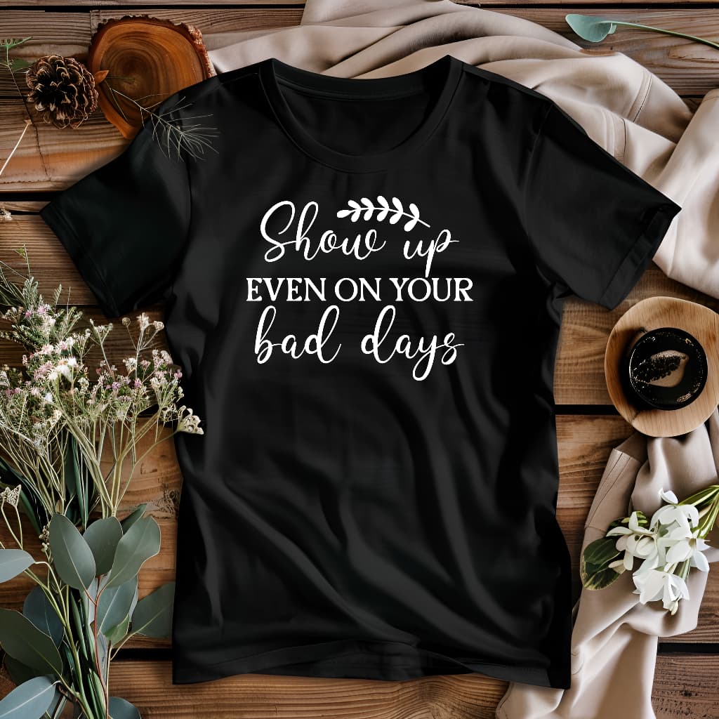 Black t-shirt with 'Show up even on your bad days' text on a wooden surface with decorative items.