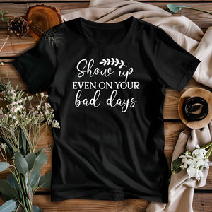 Black t-shirt with 'Show up even on your bad days' text on a wooden surface with decorative items.
