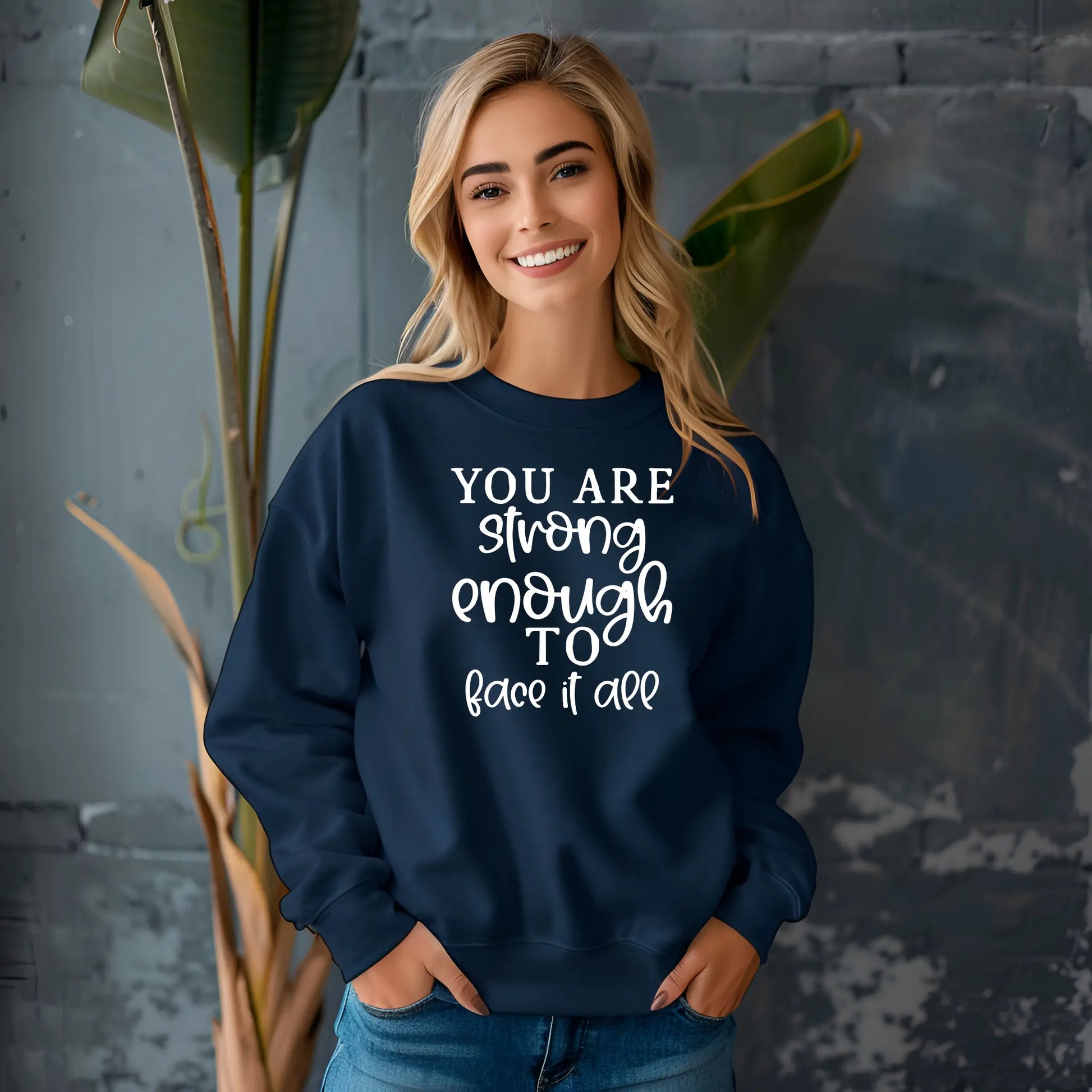 Woman wearing a navy blue sweatshirt with motivational text, standing against a gray wall with plants.
