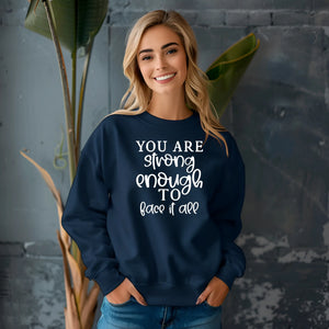 Woman wearing a navy blue sweatshirt with motivational text, standing against a gray wall with plants.