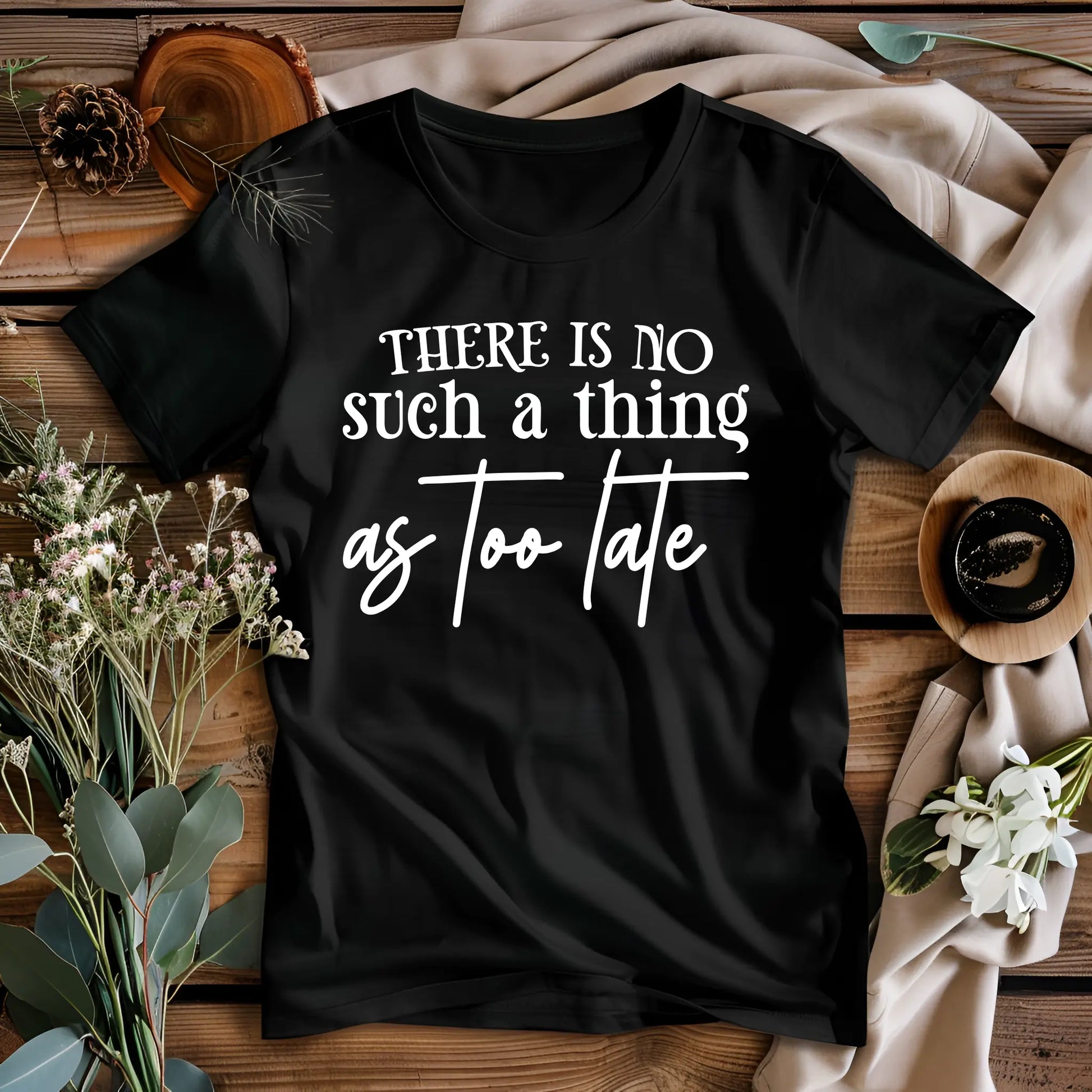 Black t-shirt with 'There is no such a thing as too late' text on a wooden surface with flowers and pinecones.