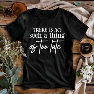 Black t-shirt with 'There is no such a thing as too late' text on a wooden surface with flowers and pinecones.